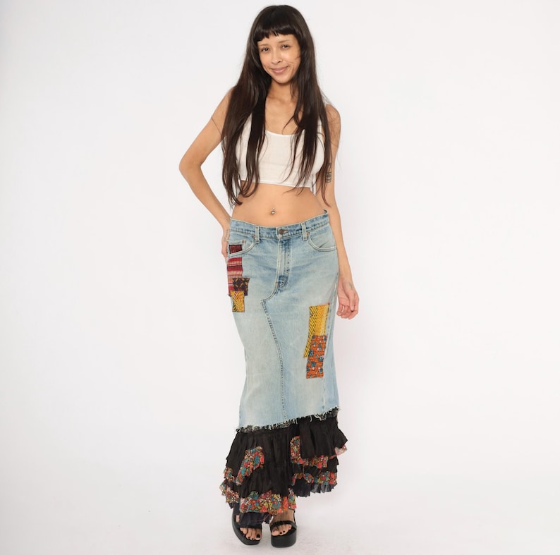 May include: A light blue denim skirt with colourful patchwork details and a black ruffled hem. The skirt is worn with a white crop top and black platform sandals. The skirt has a long, flowing silhouette.