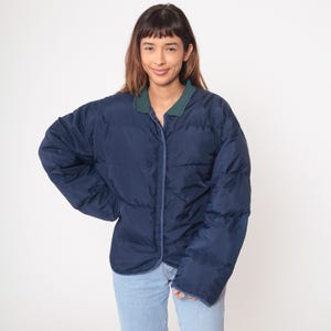 Puede incluir: Chaqueta acolchada azul marino con cuello verde. La chaqueta tiene cierre de botones en la parte delantera y un diseño acolchado. Las mangas son largas y la chaqueta parece de gran tamaño. La chaqueta se combina con vaqueros azul claro.
