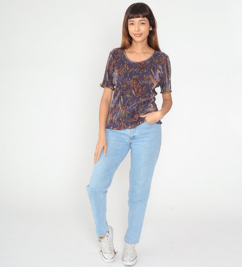 Puede incluir: Una mujer lleva una blusa de color morado y dorado con estampado floral, cuello redondo y mangas cortas. Tambi&eacute;n lleva pantalones vaqueros de color azul claro y zapatillas blancas.