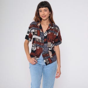 May include: Short-sleeved shirt with a geometric pattern in brown, black, and white. The shirt has a V-neck collar and is worn with light blue jeans. A casual, vintage-inspired top, suitable for everyday wear.