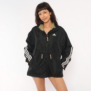 May include: A black windbreaker jacket featuring a white Adidas logo on the chest, three white stripes along each sleeve, and olive green accents on the inner collar and zipper pulls. The jacket has a full-length front zipper and two front pockets.