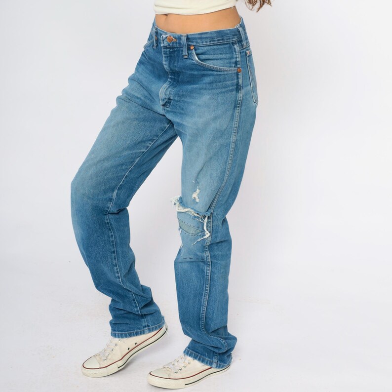 May include: Blue denim jeans with a distressed knee detail. The jeans have a classic five-pocket design and a relaxed fit. The model is wearing white trainers.