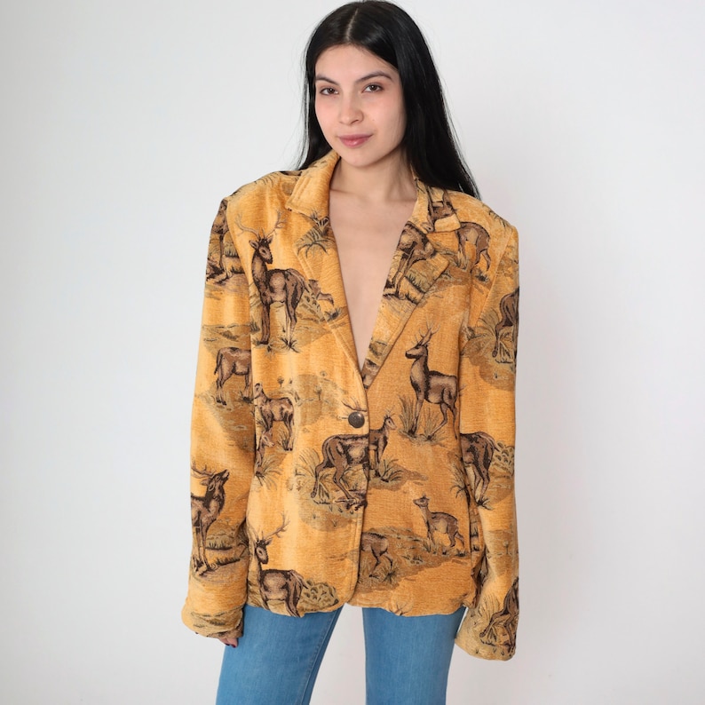 May include: A mustard-colored blazer with a deer pattern. The blazer has a single button closure and notched lapels. The deer pattern is in shades of brown and black, set against a mustard background. The blazer is worn over blue jeans.