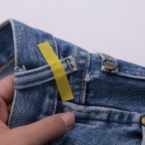 May include: Close-up of a pair of blue denim jeans with a yellow tag attached to the waistband. The jeans have a button fly and a classic five-pocket design.