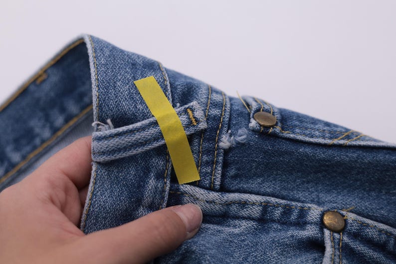May include: Close-up of a pair of blue denim jeans with a yellow tag attached to the waistband. The jeans have a button fly and a classic five-pocket design.