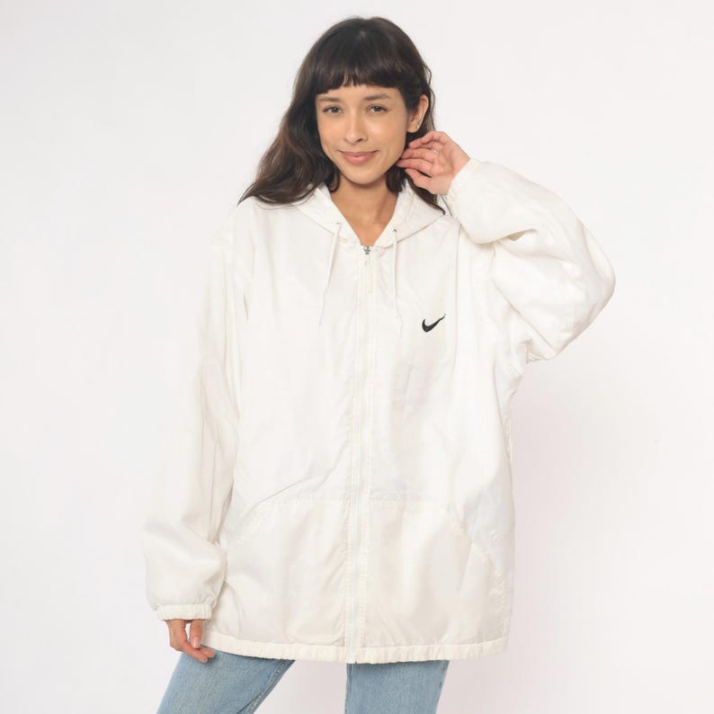 Puede incluir: Chaqueta con capucha blanca Nike con el logotipo negro de Nike en el pecho. La chaqueta tiene cierre de cremallera y capucha con cord&oacute;n. La chaqueta la lleva una persona.