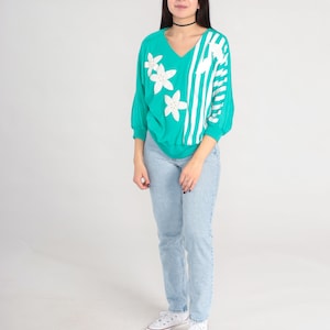 May include: A turquoise sweater with white stripes and white flower appliques. The sweater has a v-neck and long sleeves. The model is wearing light blue jeans and white sneakers.