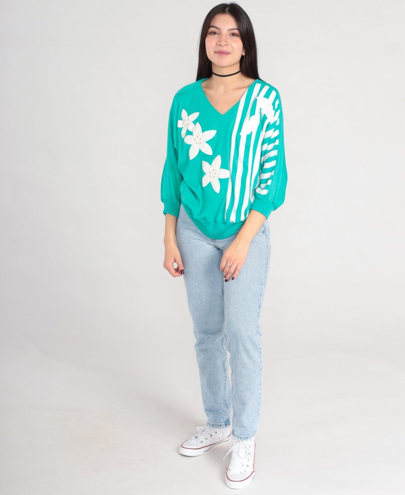 May include: A turquoise sweater with white stripes and white flower appliques. The sweater has a v-neck and long sleeves. The model is wearing light blue jeans and white sneakers.