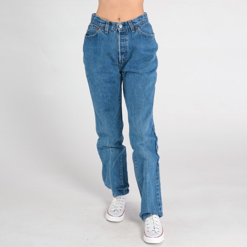 May include: A pair of blue denim jeans with a high waist and straight legs. The jeans have a button fly and five pockets. The model is wearing white trainers.