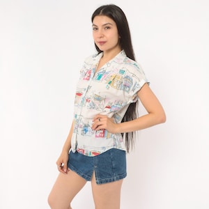 May include: A short-sleeved button-down shirt in off-white featuring a colorful hand-drawn style print of various buildings and architectural structures in shades of blue, red, yellow, and green, paired with a dark blue denim mini skirt with a front slit.