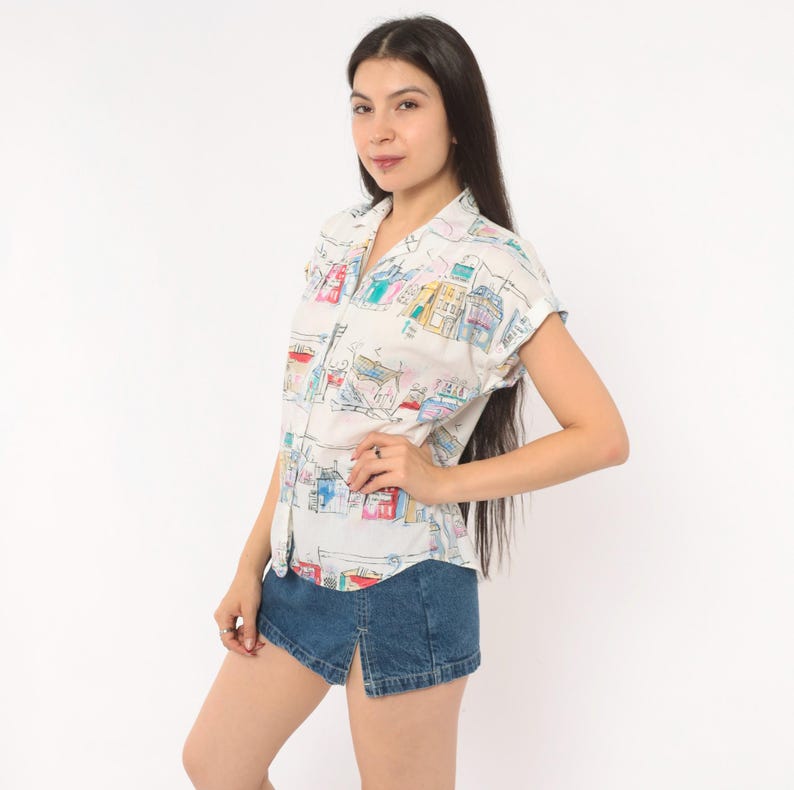 May include: A short-sleeved button-down shirt in off-white featuring a colorful hand-drawn style print of various buildings and architectural structures in shades of blue, red, yellow, and green, paired with a dark blue denim mini skirt with a front slit.