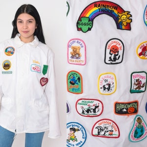 May include: A white jacket adorned with a variety of colourful embroidered patches. The patches depict various themes, including animals, holidays, and activities. The text on the patches includes "Rainbow Troop", "Teddy Bear Tea Party", "Halloween", "Fire Tour", "Hiking", "Calendar", "Holiday Parade", "Gorgonip Council", "Outdoor Cook", "Elsinore Valley Dog Test", "Skating", "Expedition Award", "Antarctica Sea World", and "Solo Fest".