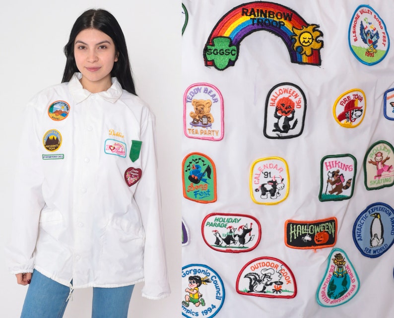 Op de afbeelding: Een wit jack met een verscheidenheid aan kleurrijke geborduurde patches. De patches tonen verschillende thema's, waaronder dieren, feestdagen en activiteiten. De tekst op de patches omvat "Rainbow Troop", "Teddy Bear Tea Party", "Halloween", "Fire Tour", "Hiking", "Calendar", "Holiday Parade", "Gorgonip Council", "Outdoor Cook", "Elsinore Valley Dog Test", "Skating", "Expedition Award", "Antarctica Sea World" en "Solo Fest".