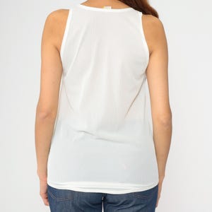 Peut inclure: D&eacute;bardeur blanc sans manches avec un col rond. Le haut est fait d'un tissu l&eacute;ger et l&eacute;g&egrave;rement transparent. Le dos du haut est visible, montrant la coupe et la coupe. Le mod&egrave;le porte un jean bleu.