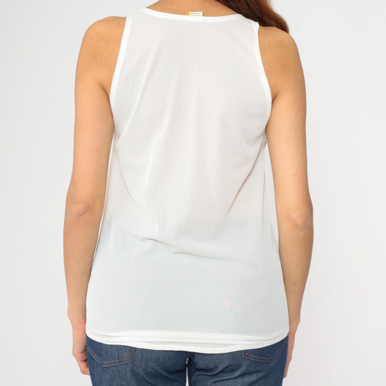 Peut inclure: D&eacute;bardeur blanc sans manches avec un col rond. Le haut est fait d'un tissu l&eacute;ger et l&eacute;g&egrave;rement transparent. Le dos du haut est visible, montrant la coupe et la coupe. Le mod&egrave;le porte un jean bleu.