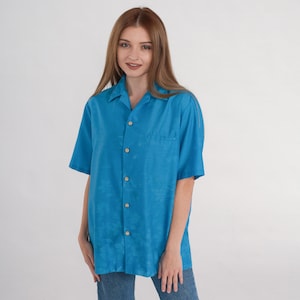 May include: A short-sleeved, turquoise button-down shirt with a chest pocket. The shirt has a classic collar and wooden buttons. The fabric has a subtle textured pattern. The shirt is paired with blue jeans.