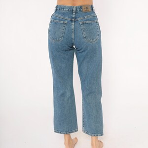 May include: Light blue denim jeans with a high waist and straight leg. The jeans have classic five-pocket styling and a leather patch on the back waistband. The jeans are a medium wash and appear to be vintage.