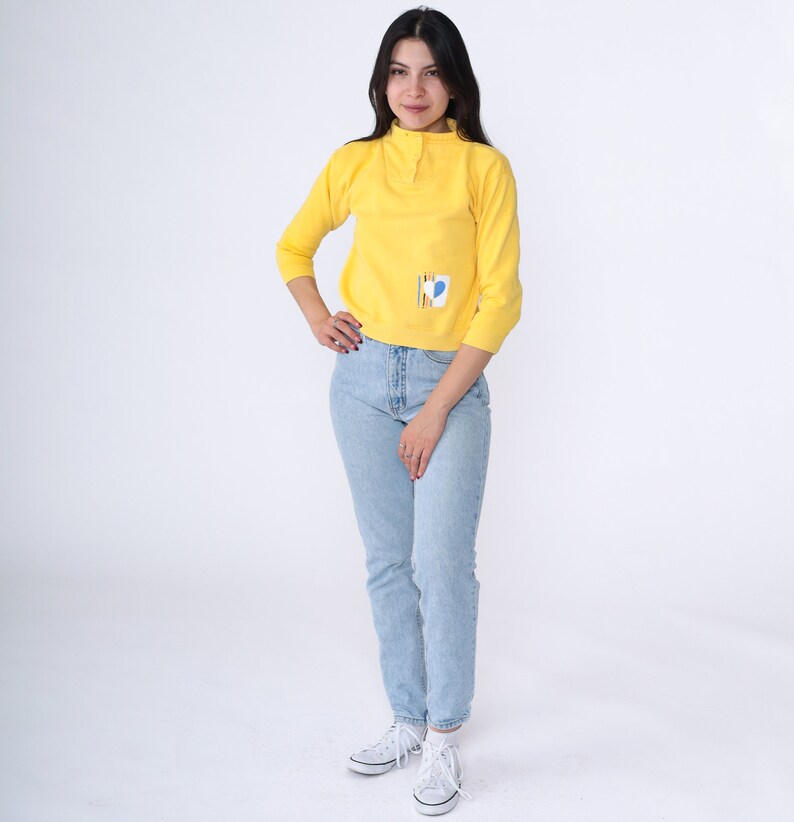 May include: A woman wearing a yellow long-sleeved shirt with a geometric design on the front and light blue jeans. She is standing on a white background.