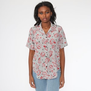 May include: Short-sleeved button-down shirt with a gray background and a repeating pattern of red and black abstract shapes. The shirt has a collared neckline and black buttons.