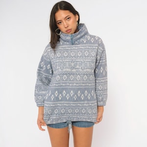 May include: A light blue fleece pullover featuring a white geometric and snowflake pattern. The garment has a high collar with a partial front zipper closure, long sleeves with elastic cuffs, and an adjustable drawstring hem at the waist.