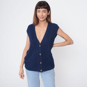 May include: A navy blue, sleeveless cardigan with a V-neck and wooden buttons. The knitted sweater has a cable knit pattern and is worn with light blue jeans. The cardigan is a classic style, suitable for layering.