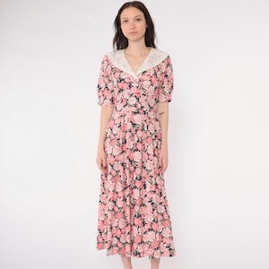 Op de afbeelding: Een vintage bloemen midi-jurk met een witte kraag. De jurk heeft een roze en marine bloemenpatroon, korte mouwen en een knoopsluiting aan de voorkant. De jurk is knielang en heeft een getailleerd lijfje.