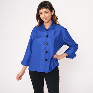 May include: A vibrant blue button-down shirt with three-quarter sleeves and a classic collar. The shirt features large black buttons and is made of a shiny, silky material. The garment is paired with black pants.