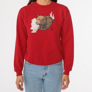 May include: Red long-sleeved sweatshirt with a brown bear graphic on the front. The bear is peeking out from behind a white cloud-like design. The sweatshirt has a crew neck and long sleeves.