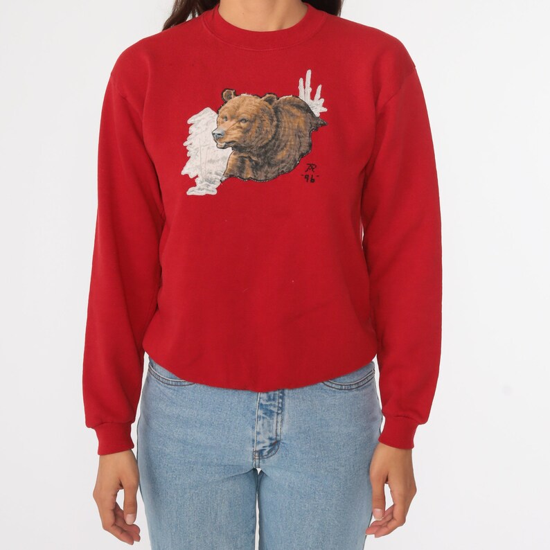May include: Red long-sleeved sweatshirt with a brown bear graphic on the front. The bear is peeking out from behind a white cloud-like design. The sweatshirt has a crew neck and long sleeves.