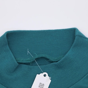 Pode incluir: Close-up de uma camisola de gola alta verde-azulado. A gola canelada &eacute; vis&iacute;vel, juntamente com uma parte do corpo da pe&ccedil;a. Uma etiqueta branca com um c&oacute;digo QR pende da gola.