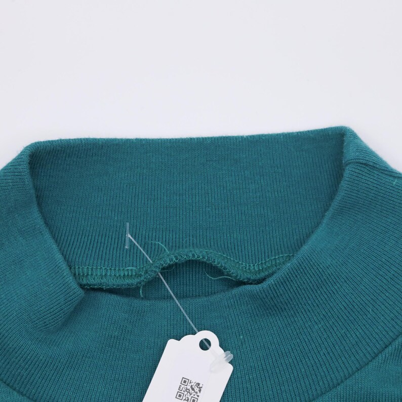 Pode incluir: Close-up de uma camisola de gola alta verde-azulado. A gola canelada &eacute; vis&iacute;vel, juntamente com uma parte do corpo da pe&ccedil;a. Uma etiqueta branca com um c&oacute;digo QR pende da gola.