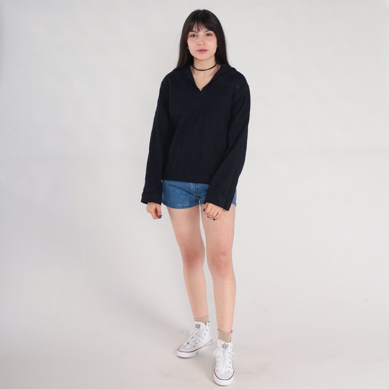 May include: A woman wearing a black long-sleeved shirt with a collar and a v-neck. She is wearing denim shorts and white sneakers.