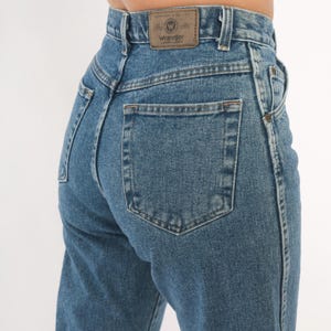 May include: Close-up of blue denim jeans with a Wrangler brand patch on the back waistband. The jeans have a classic five-pocket design and a straight leg cut. The denim is a medium wash with visible stitching.