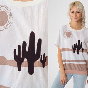 May include: White cotton t-shirt with a brown and black embroidered design of a desert landscape with a sun and cacti.