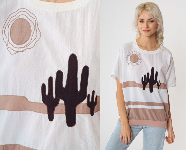 May include: White cotton t-shirt with a brown and black embroidered design of a desert landscape with a sun and cacti.