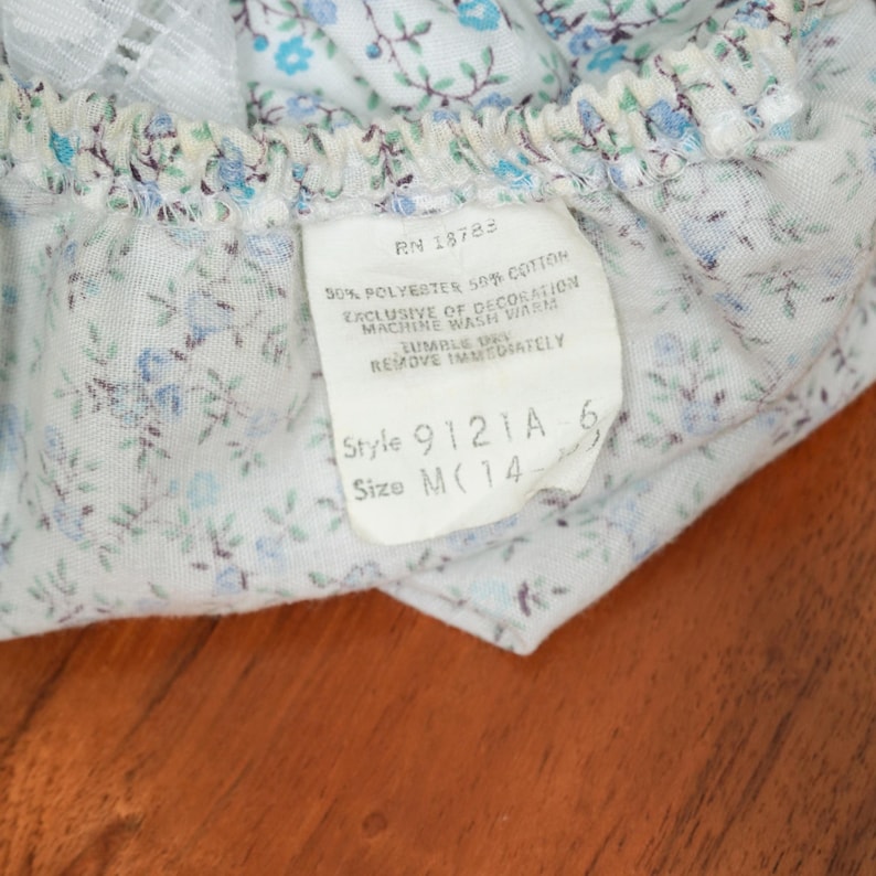 May include: A white cotton and polyester fabric with a blue and green floral print. The fabric has a tag attached with the text "RN 18783 50% POLYESTER 50% COTTON EXCLUSIVE OF DECORATION MACHINE WASH WARM TUMBLE REMOVE IMMEDIATELY Style 9121A 6 Size M(14"