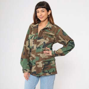 May include: A green, brown, and black camouflage jacket with a button closure and two large pockets. The jacket has "U.S. ARMY" embroidered on the left chest.