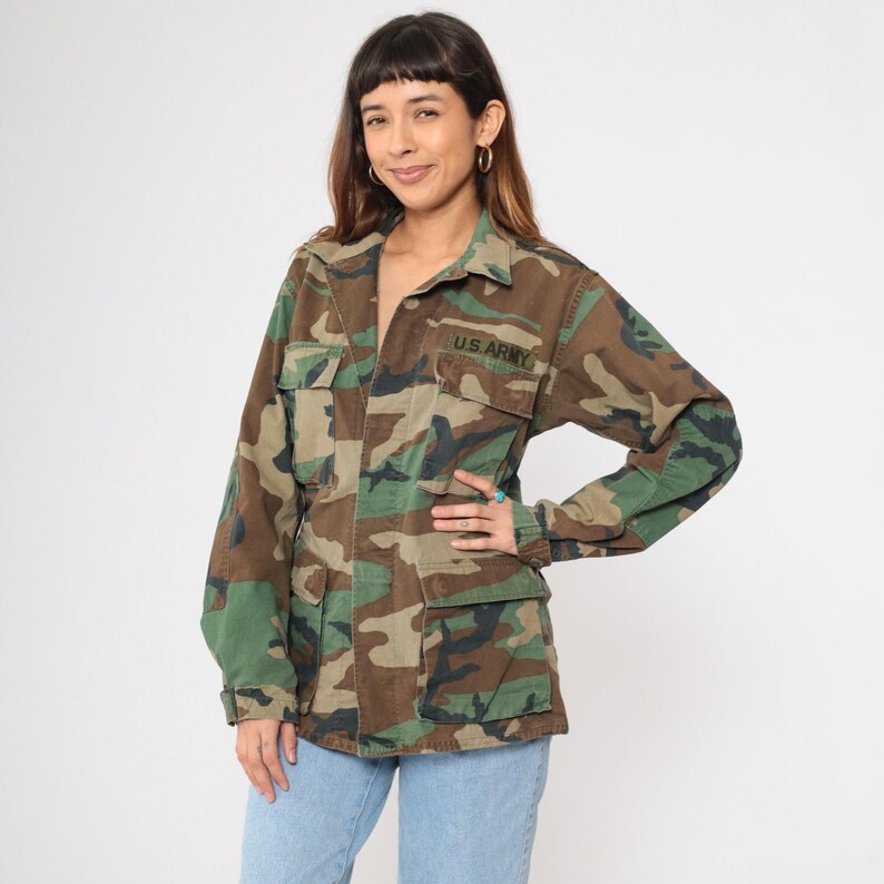 May include: A green, brown, and black camouflage jacket with a button closure and two large pockets. The jacket has "U.S. ARMY" embroidered on the left chest.