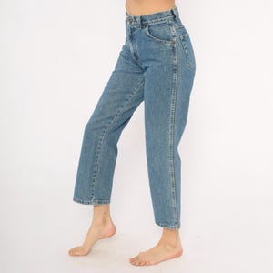 May include: Light blue denim jeans with a classic five-pocket design. The jeans have a straight leg cut and a high waist. The denim is a medium wash, and the jeans are worn at the ankles. The jeans are a casual style.