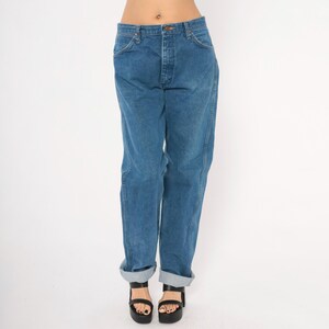 May include: Blue denim jeans with a button fly and classic five-pocket design. The jeans have a relaxed fit and are cuffed at the ankles. The model is wearing black platform sandals.