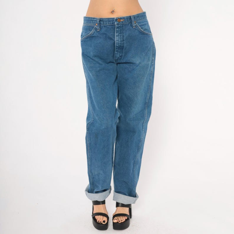 May include: Blue denim jeans with a button fly and classic five-pocket design. The jeans have a relaxed fit and are cuffed at the ankles. The model is wearing black platform sandals.