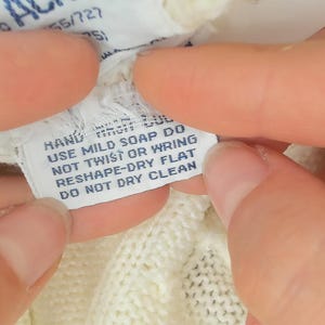 May include: Close-up of a white clothing tag with care instructions. The tag reads "HAND USE MILD SOAP DO NOT TWIST OR WRING RESHAPE-DRY FLAT DO NOT DRY CLEAN". The tag is held between two hands.