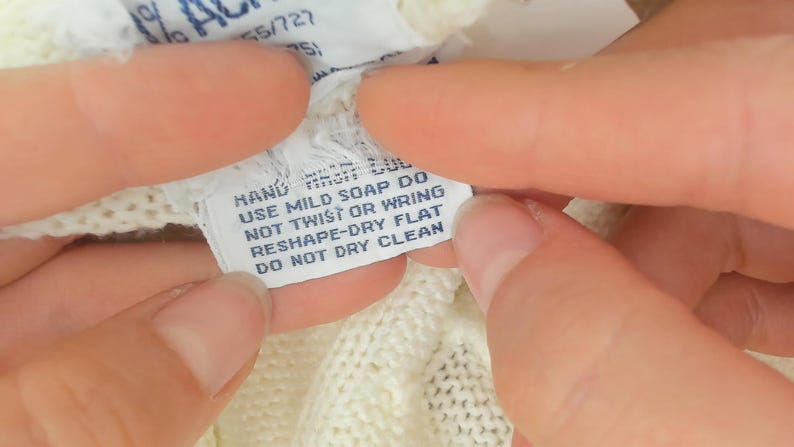 May include: Close-up of a white clothing tag with care instructions. The tag reads "HAND USE MILD SOAP DO NOT TWIST OR WRING RESHAPE-DRY FLAT DO NOT DRY CLEAN". The tag is held between two hands.