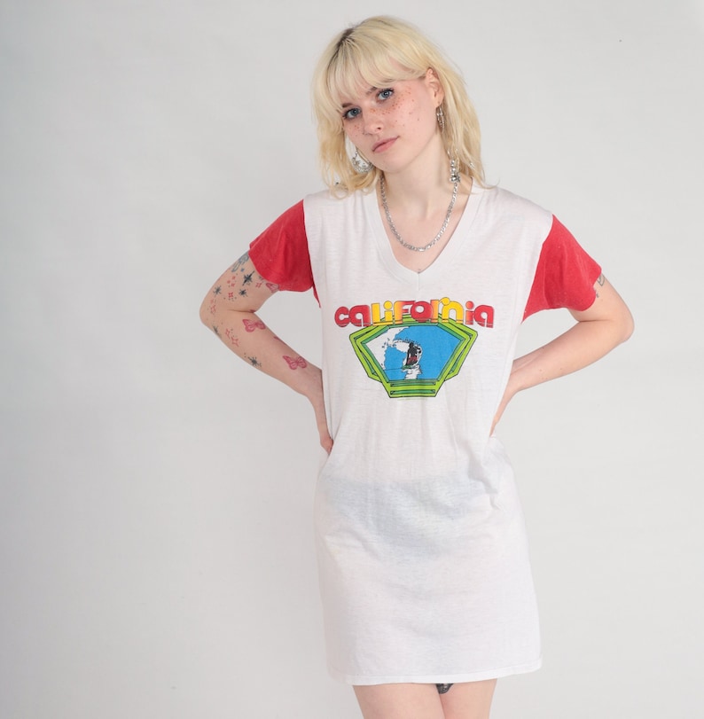 K&ouml;nnte beinhalten: Wei&szlig;es T-Shirt-Kleid mit roten &Auml;rmeln und einem Grafikdruck eines Surfers mit dem Wort "California" in einer bunten Schrift.