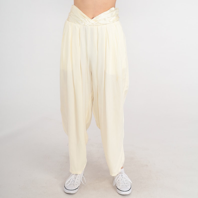 May include: Cream-colored harem pants with a high, twisted waistband. The pants have a loose, flowing fit and are paired with white sneakers. The fabric appears to be lightweight and slightly sheer, with vertical pleats adding texture.