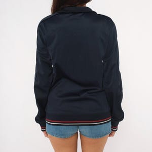 May include: A dark navy blue long-sleeve track jacket featuring a high collar and a full-length front zipper. The hem and sleeve cuffs are finished with a ribbed knit trim in contrasting white and red stripes against a dark navy blue background.
