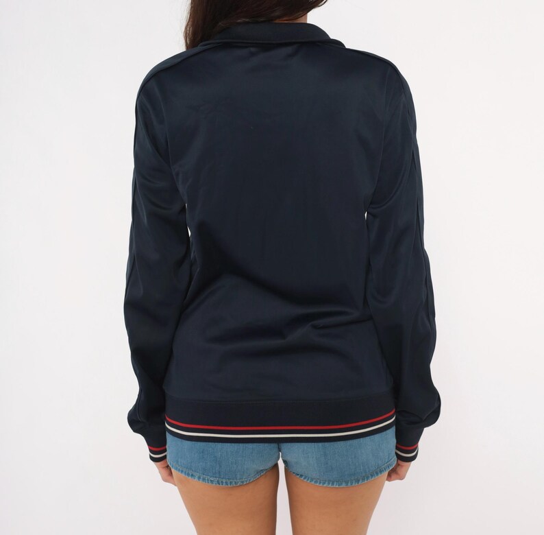 May include: A dark navy blue long-sleeve track jacket featuring a high collar and a full-length front zipper. The hem and sleeve cuffs are finished with a ribbed knit trim in contrasting white and red stripes against a dark navy blue background.