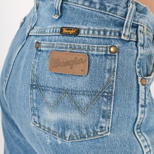 Pu&ograve; includere: Primo piano di jeans di denim blu chiaro. La tasca posteriore presenta un classico design a punto 'W' e una toppa in pelle marrone con il logo 'Wrangler'. I jeans hanno rivetti color ottone e una piccola etichetta nera con il marchio 'Wrangler'.