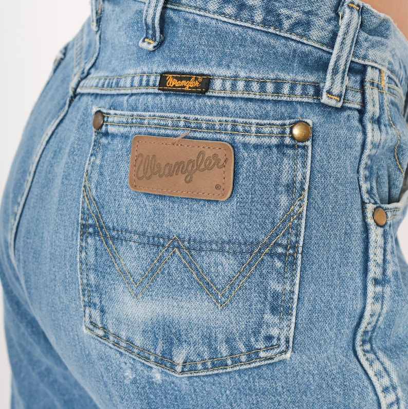 Pu&ograve; includere: Primo piano di jeans di denim blu chiaro. La tasca posteriore presenta un classico design a punto 'W' e una toppa in pelle marrone con il logo 'Wrangler'. I jeans hanno rivetti color ottone e una piccola etichetta nera con il marchio 'Wrangler'.