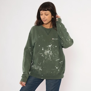Puede incluir: Una sudadera Champion verde oliva con salpicaduras de pintura blanca. La sudadera tiene cuello redondo y el logo de Champion bordado en el pecho. La modelo lleva vaqueros azules.